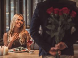 Beautiful girl is sitting at the table in restaurant and smiling while a man is holding roses behind his back