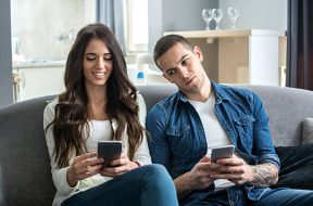 Young couple using smartphones at home. Jealous boyfriend spying his girlfriend's phone.