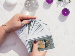 Hands with purple nails and rings hold deck of Tarot cards on white surface with crystal ball, candles and stones. Top view. Minsk, Belarus - 28.07.2021