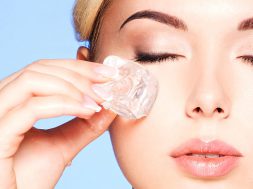 Closeup portrait of beautiful young woman applies the ice to face near eyes on a blue background.