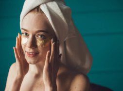 Caucasian freckled woman applying golden collagen patches under her eyes, looking at camera. Natural beauty. Facial treatment.