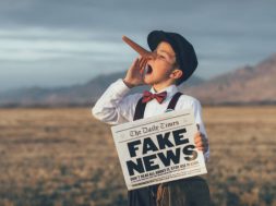 A news boy dressed in vintage knickers, newsboy hat and fake long Pinocchio nose stands with a fake newspaper in the middle of a field in Utah, USA. He is trying to sell you fake news.