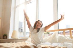 Portrait of beautiful Mixed-Race woman stretching sitting in bed lit by sunlight, copy space