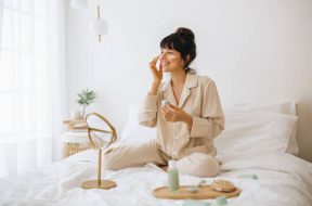 Happy woman doing routine skin care at home with beauty products. Woman sitting on bed at home and applying face cream.