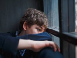 Pensive sad boy teenager with blue eyes in a blue shirt and jeans sitting at the window and closes his face with his hands.